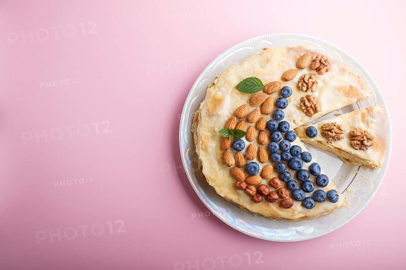 Homemade layered Napoleon cake with milk cream. Decorated with blueberry, almonds, walnuts, hazelnuts, mint on a pastel pink background. top view. flat lay, copy space