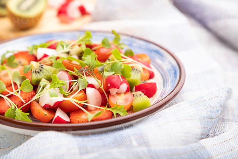 Vegetarian fruits and vegetables salad of strawberry, kiwi, tomatoes, microgreen sprouts on white concrete background and linen textile. Side view, close up, selective focus