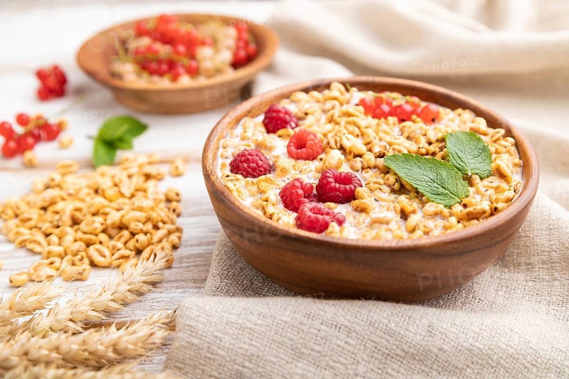 Wheat flakes porridge with milk, raspberry and currant in wooden bowl on white wooden background and linen textile. Side view, close up, selective focus