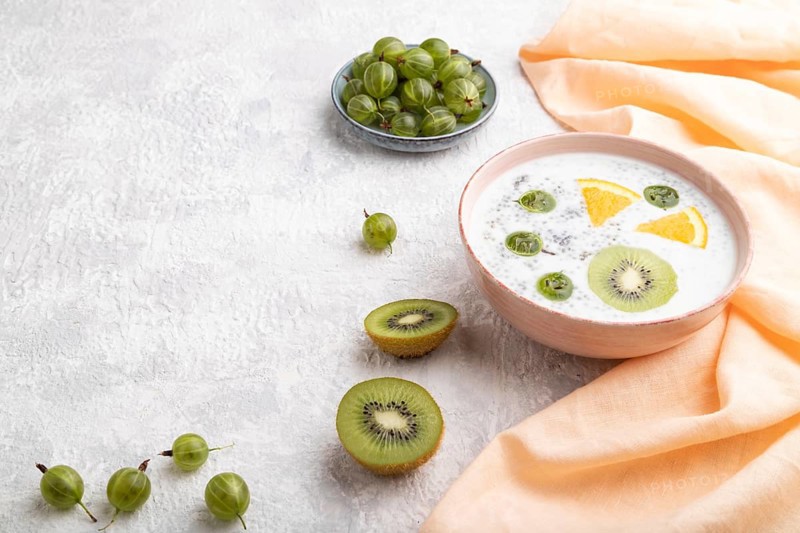 Yogurt with kiwi, gooseberry, chia in ceramic bowl on gray concrete background and orange linen textile. Side view, copy space