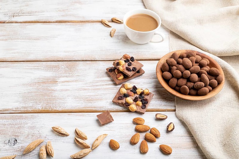 Almond in chocolate dragees in wooden plate and a cup of coffee on white wooden background and linen textile. Side view, copy space