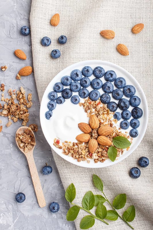 Yoghurt with blueberry, granola and almond in white plate with wooden spoon on gray concrete background and linen textile. top view, flat lay, close up