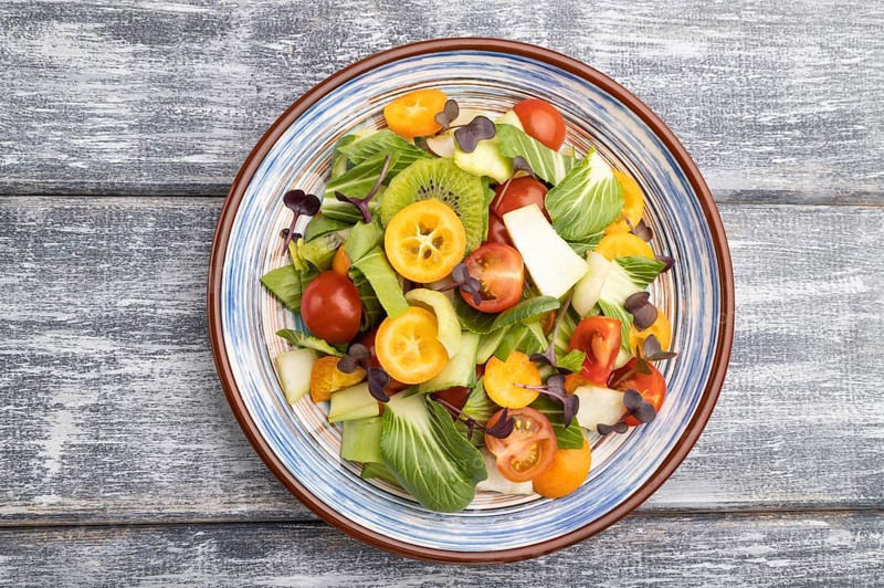 Vegetarian salad of pac choi cabbage, kiwi, tomatoes, kumquat, microgreen sprouts on gray wooden background. Top view, flat lay, close up