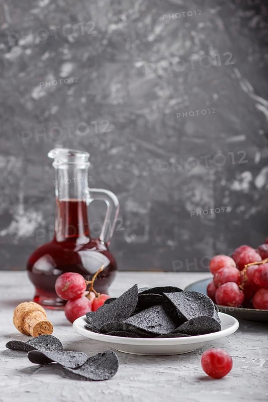 Black potato chips with charcoal, balsamic vinegar in glass, red grapes on a blue ceramic plate on a gray concrete background. side view, close up, selective focus, copy space