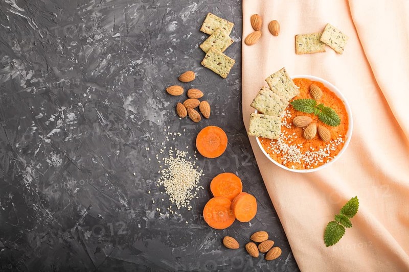 Carrot cream soup with sesame seeds, almonds and snacks in white bowl on a black concrete background with orange textile. top view, flat lay, copy space