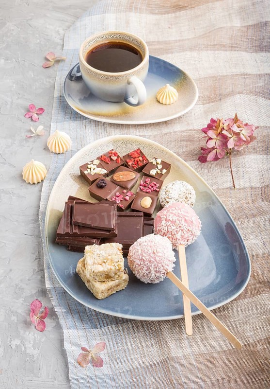 A pieces of homemade chocolate with coconut candies and a cup of coffee on a gray concrete background and blue and brown textile. side view, close up