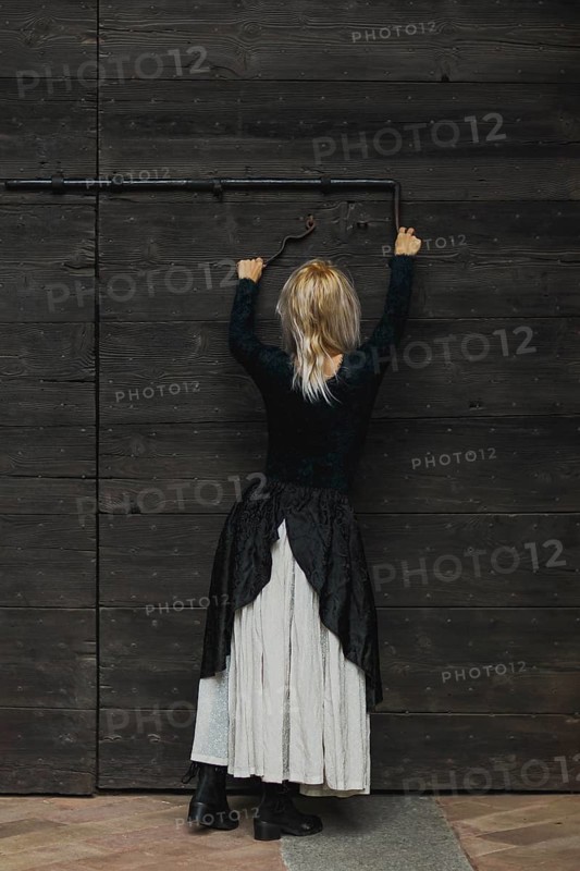 Full body shot of a young blonde caucasian woman reaching an antique lock of a wooden door above her head, wearing a black top and white skirt and a furry shirt