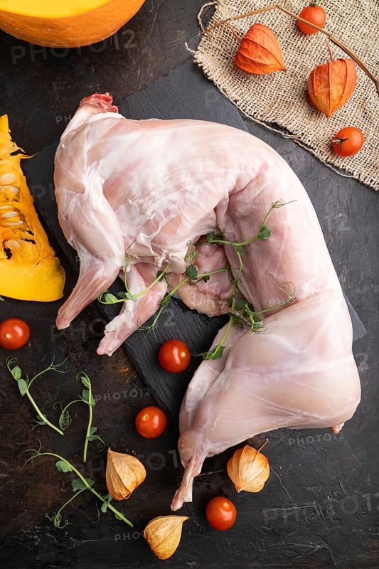Whole raw rabbit with pumpkin, tomatoes, pea sprouts on a black concrete background and linen textile. Top view, flat lay, close up