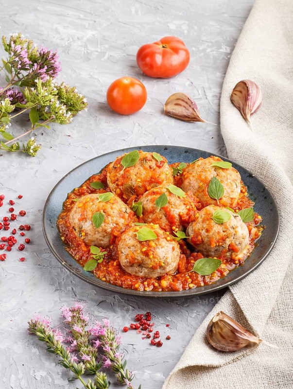 Pork meatballs with tomato sauce, oregano leaves, spices and herbs on blue ceramic plate on a gray concrete background with linen textile. side view, close up