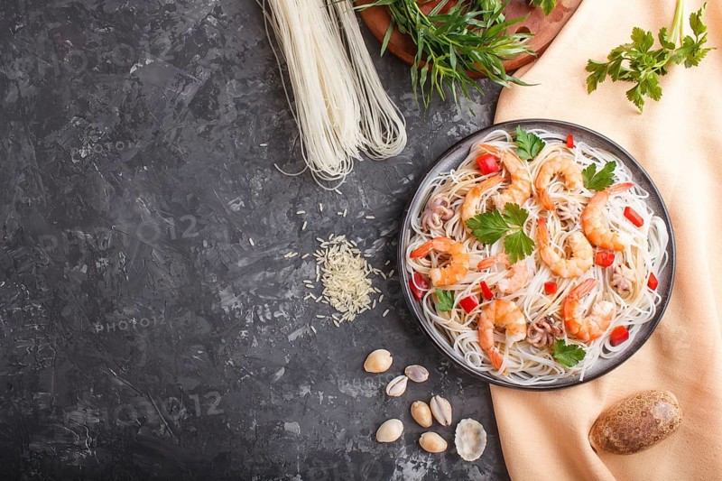 Rice noodles with shrimps or prawns and small octopuses on gray ceramic plate on a black concrete background and orange textile. Top view, flat lay, copy space