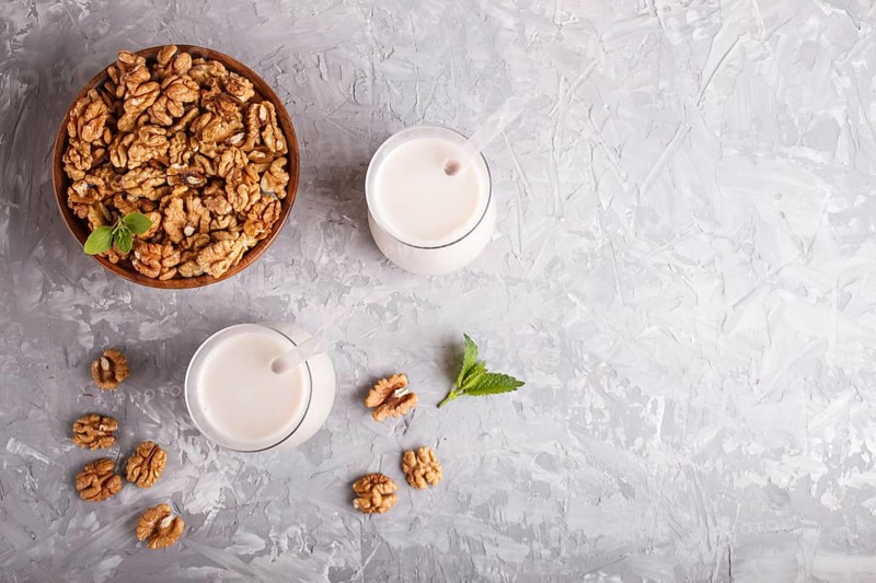 Organic non dairy walnut milk in glass and wooden plate with walnuts on a gray concrete background. Vegan healthy food concept, flat lay, top view, copy space