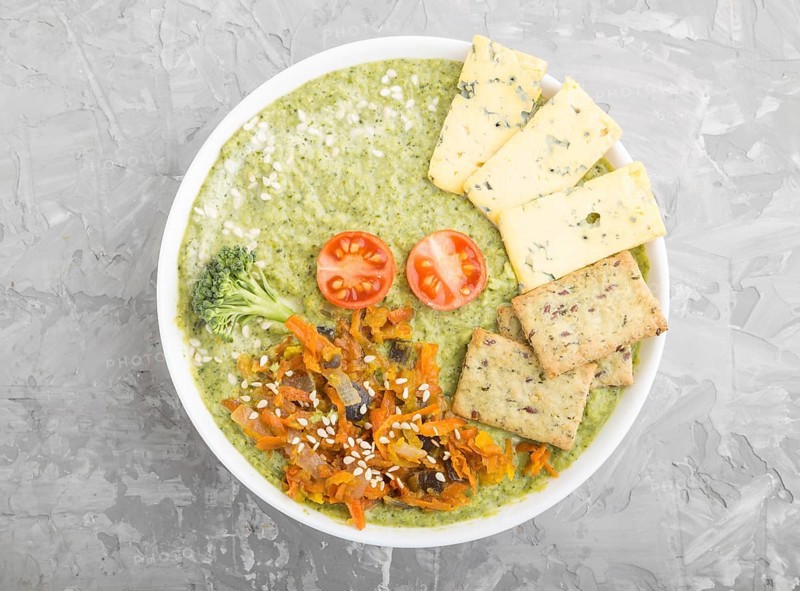 Green broccoli cream soup with crackers and cheese in white bowl on a gray concrete background. top view, flat lay, close up