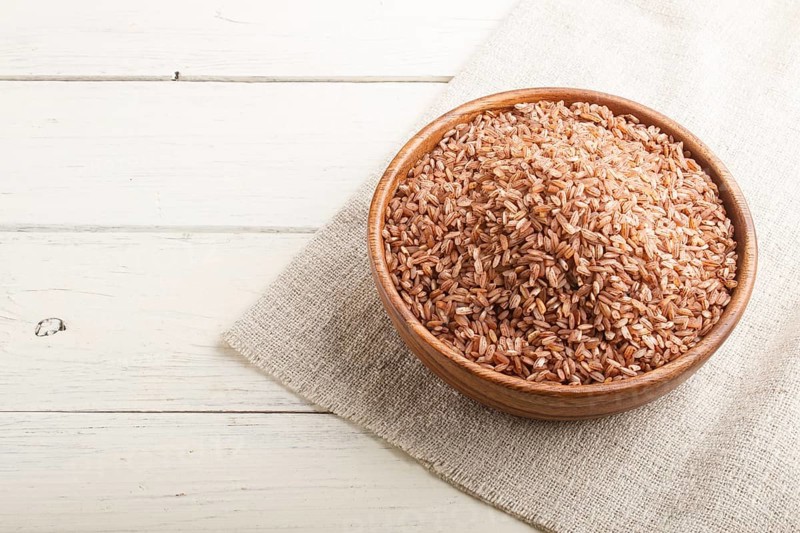 Wooden bowl with unpolished brown rice on a white wooden background and linen textile. Side view, copy space