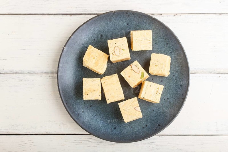 Traditional indian candy soan papdi in a blue ceramic plate with almond and pistache on a white wooden background. top view, flat lay, close up