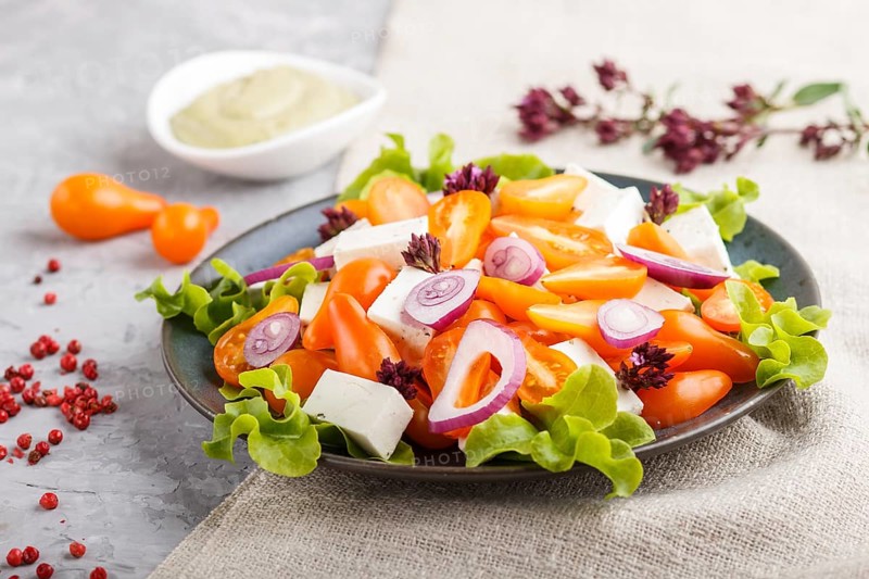 Vegetarian salad with fresh grape tomatoes, feta cheese, lettuce and onion on blue ceramic plate on gray concrete background and linen textile. side view, close up, selective focus