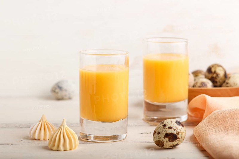 Sweet egg liqueur in glass with quail eggs and meringues on white wooden background. Side view, close up, high key
