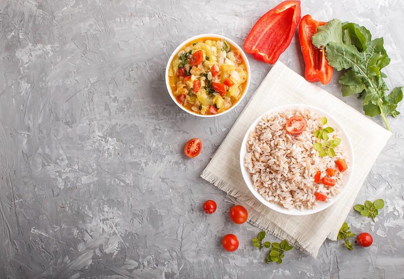 Unpolished rice porridge with stewed vegetables and oregano in white bowl on a gray concrete background and linen textile. Top view, flat lay, copy space