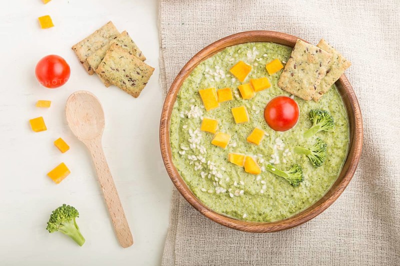 Green broccoli cream soup with crackers and cheese in wooden bowl on a white wooden background and linen napkin. top view, flat lay, close up