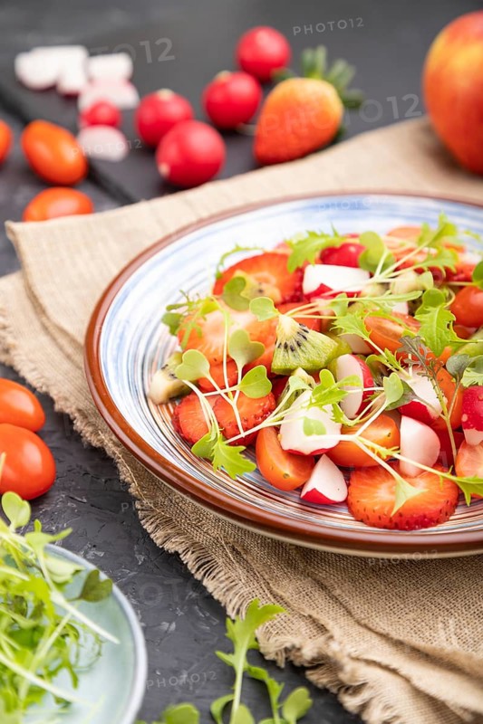 Vegetarian fruits and vegetables salad of strawberry, kiwi, tomatoes, microgreen sprouts on black concrete background and linen textile. Side view, close up, selective focus