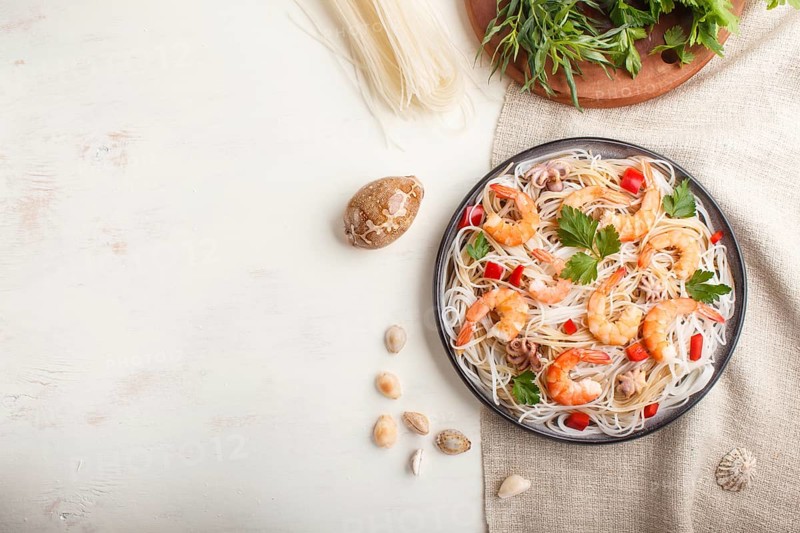 Rice noodles with shrimps or prawns and small octopuses on gray ceramic plate on a white wooden background and linen textile. Top view, flat lay, copy space