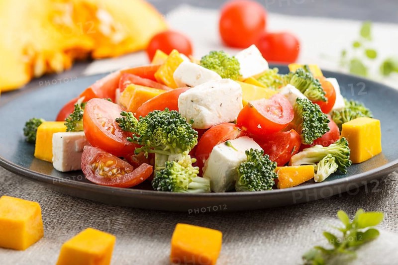 Vegetarian salad with broccoli, tomatoes, feta cheese, and pumpkin on a blue ceramic plate on a black concrete background and linen textile, side view, close up, selective focus