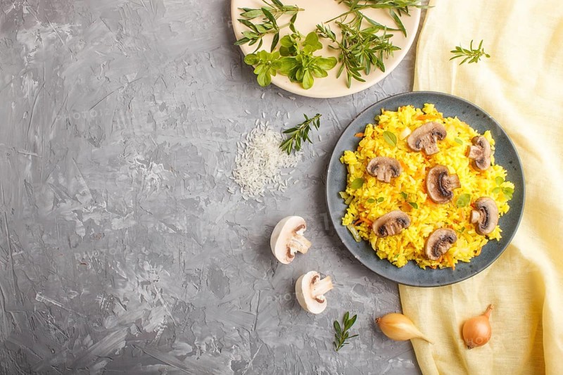 Yellow fried rice with champignons mushrooms, turmeric and oregano on blue ceramic plate on a gray concrete background and yellow textile. Top view, flat lay, copy space