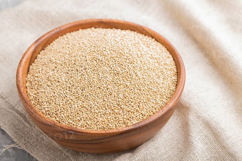Wooden bowl with raw white quinoa seeds and wooden spoon on a gray concrete background and linen textile. Side view, close up, selective focus