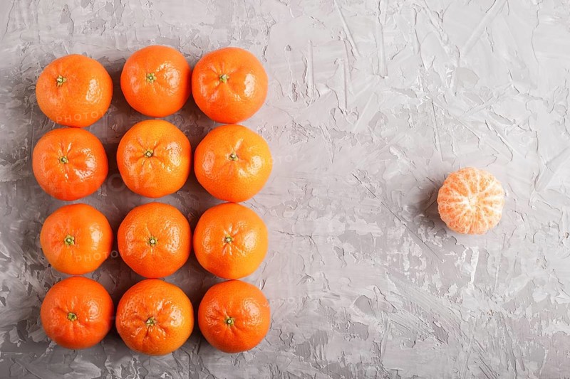 Rows of tangerines forming a rectangle and one peeled tangerine on a gray concrete background, top view, flat lay