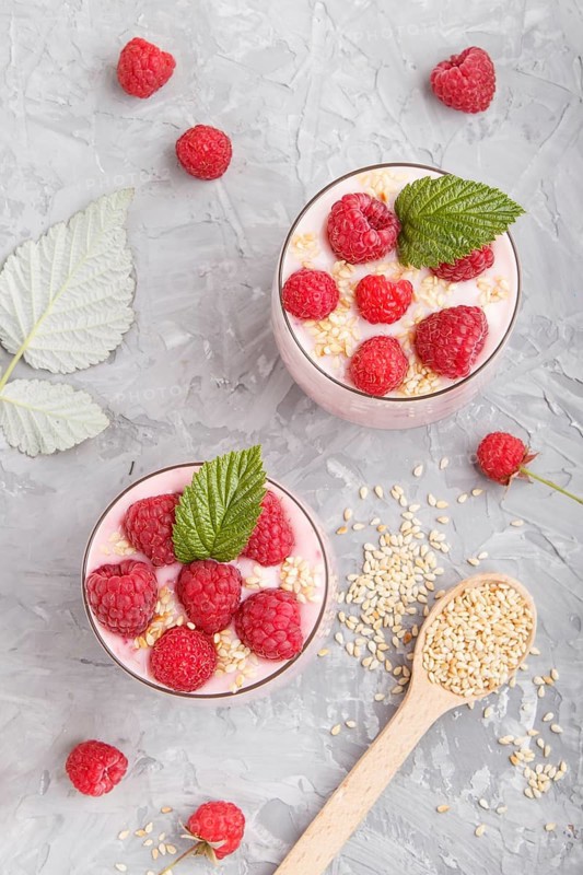 Yoghurt with raspberry and sesame in a glass and wooden spoon on gray concrete background. top view, flat lay, close up