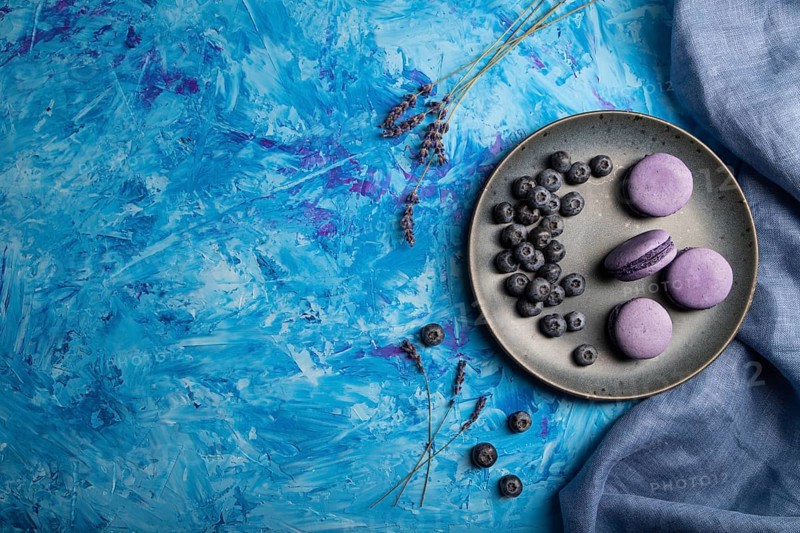 Purple macarons or macaroons cakes with blueberries on ceramic plate on a blue concrete background and blue textile. Flat lay, top view, copy space