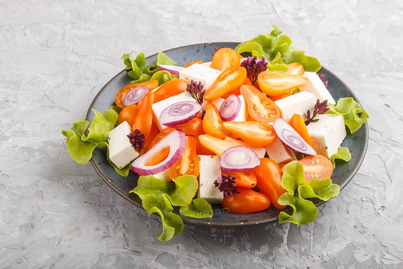 Vegetarian salad with fresh grape tomatoes, feta cheese, lettuce and onion on blue ceramic plate on gray concrete background, side view, close up