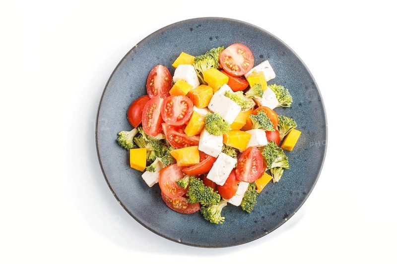 Vegetarian salad with broccoli, tomatoes, feta cheese, and pumpkin on a blue ceramic plate isolated on white background, top view, close up