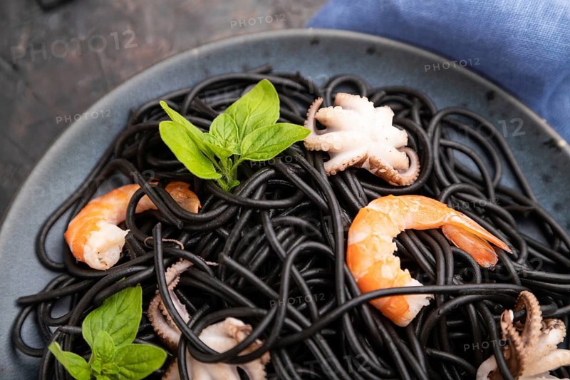 Black cuttlefish ink pasta with shrimps or prawns and small octopuses on black concrete background and blue textile. Top view, close up, selective focus
