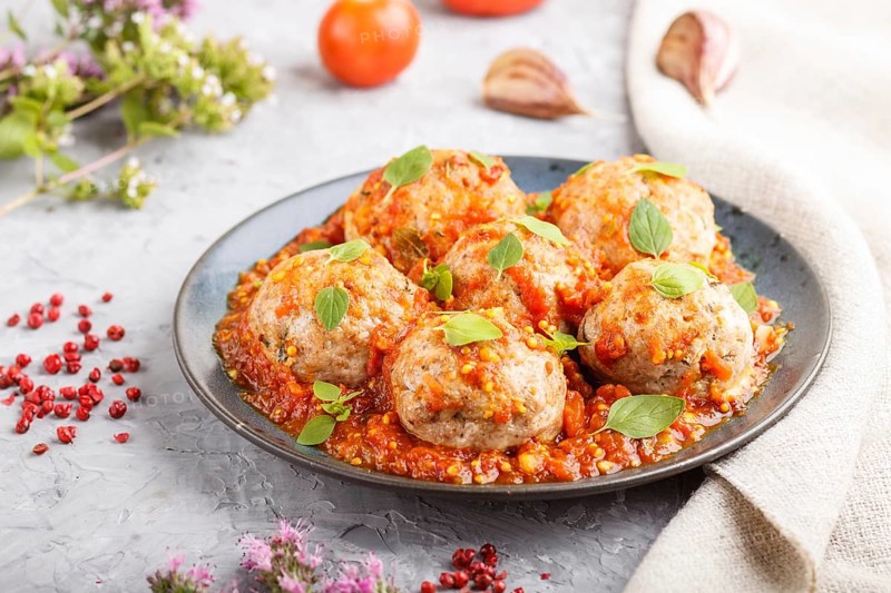 Pork meatballs with tomato sauce, oregano leaves, spices and herbs on blue ceramic plate on a gray concrete background with linen textile. side view, close up, selective focus