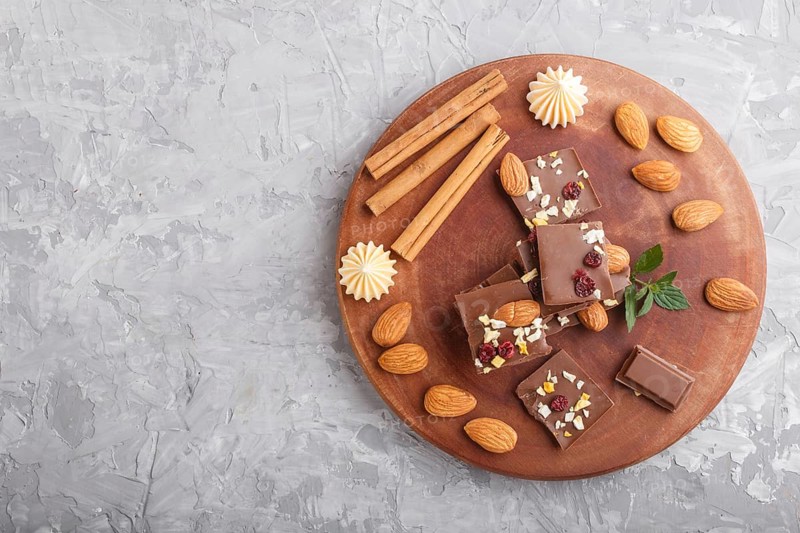 A pieces of milk chocolate with almonds and dried fruits on a brown wooden board on a gray concrete background. top view, copy space, flat lay