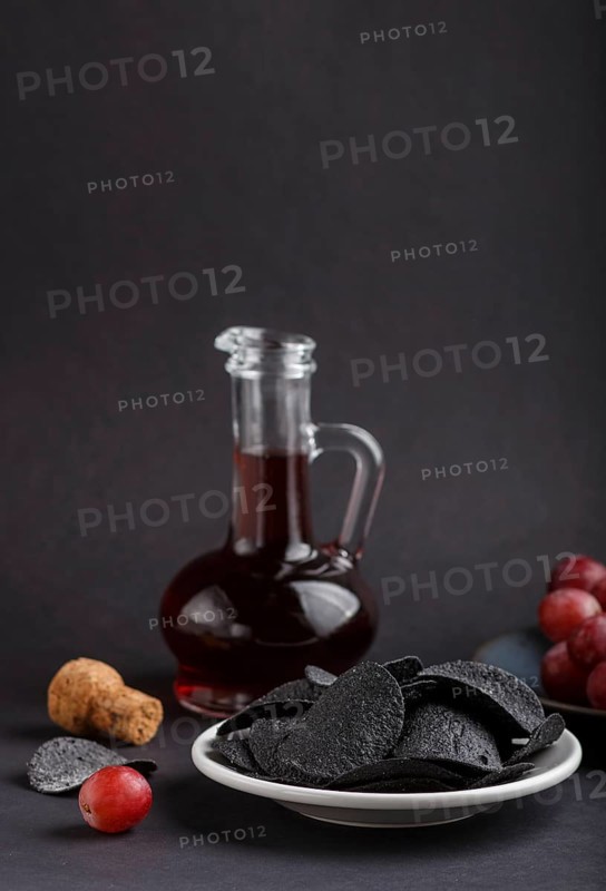 Black potato chips with charcoal, balsamic vinegar in glass, red grapes on a blue ceramic plate on a black background. side view, close up, selective focus, copy space