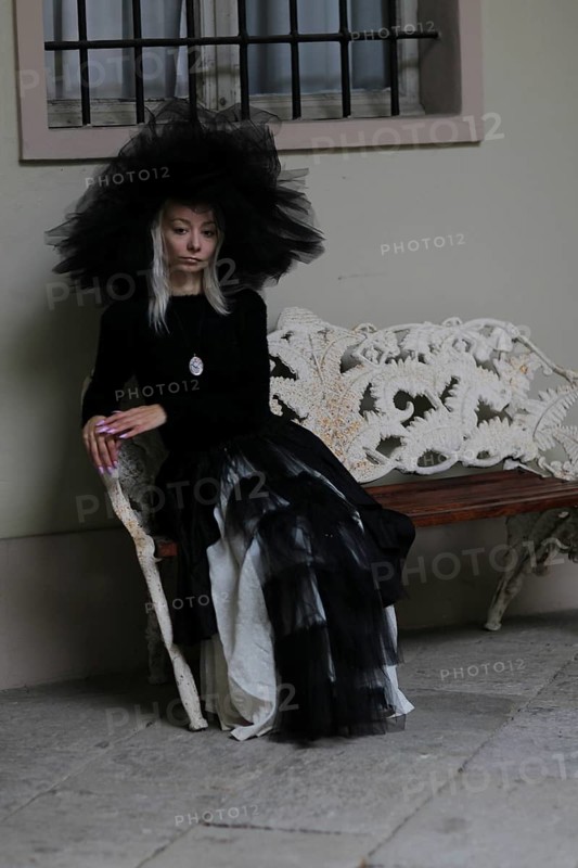 Gothically styled white thin blonde woman sitting on a lace-covered bench in a vintage indoor setting, wear lace veil and hat, black and white dress