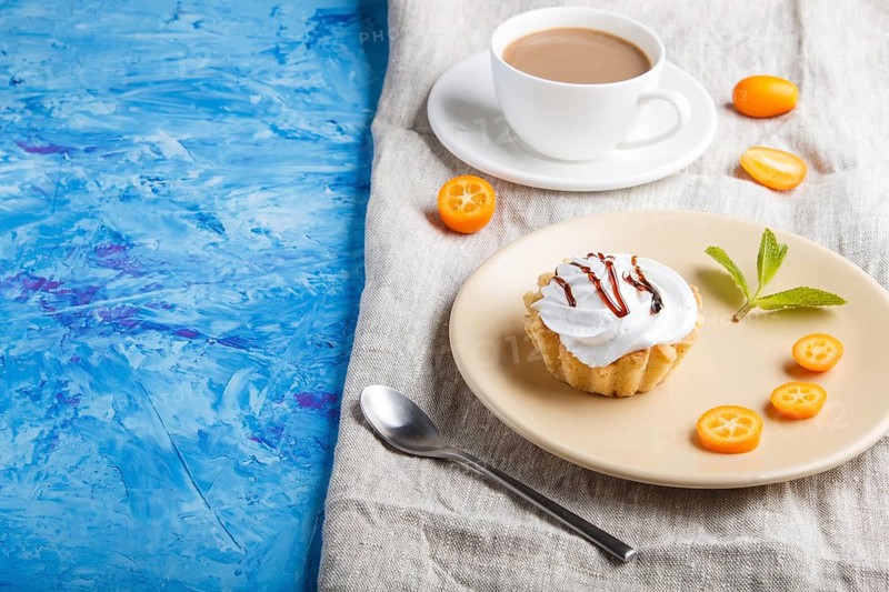 Cake with whipped egg cream on a light brown plate with kumquat slices and mint leaves on a blue concrete background with linen napkin. copy space, side view