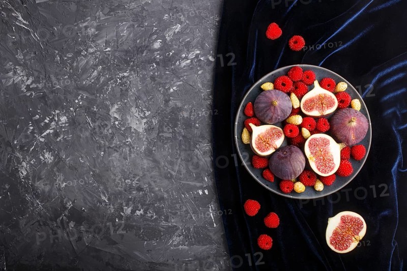 Fresh figs, strawberries and raspberries on blue ceramic plate on black concrete background and blue velvet textile. top view, flat lay, copy space