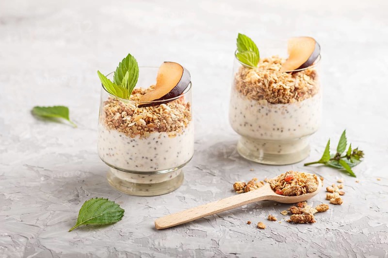 Yoghurt with plum, chia seeds and granola in a glass and wooden spoon on gray concrete background. side view, close up