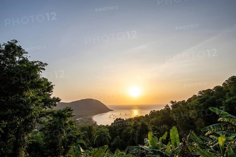 View from a mountain onto a secluded bay with a sandy beach and mangrove forest. The sun sets over the sea and boats float in the water. Grande Anse beach, Basse Terre, Guadeloupe, French Antilles, Caribbean, North America