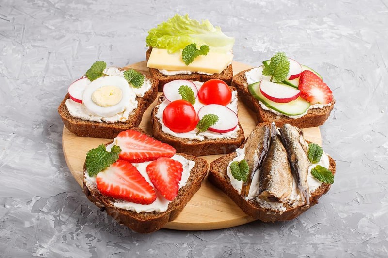 Set of different sandwiches with cheese, radish, lettuce, strawberry, sprats, tomatoes and cucumber on wooden board on a gray concrete background. side view, close up