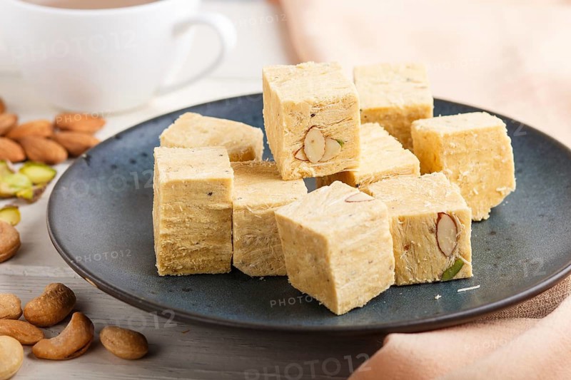 Traditional indian candy soan papdi in a blue ceramic plate with almond, pistache, cashew and a cup of coffee on a white wooden background with orange textile. side view, close up, selective focus