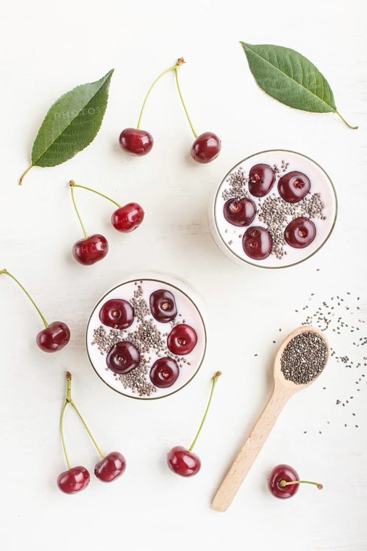 Yoghurt with cherries, chia seeds and granola in glass with wooden spoon on white wooden background. top view, flat lay