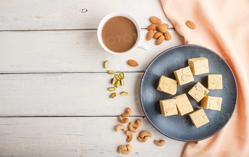 Traditional indian candy soan papdi in a blue ceramic plate with almond, pistache, cashew and a cup of coffee on a white wooden background with orange textile. top view, flat lay, copy space