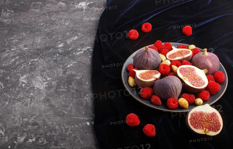 Fresh figs, strawberries and raspberries on blue ceramic plate on black concrete background and blue velvet textile. side view, copy space