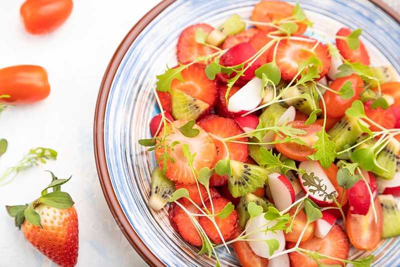 Vegetarian fruits and vegetables salad of strawberry, kiwi, tomatoes, microgreen sprouts on white concrete background and linen textile. Top view, close up, selective focus