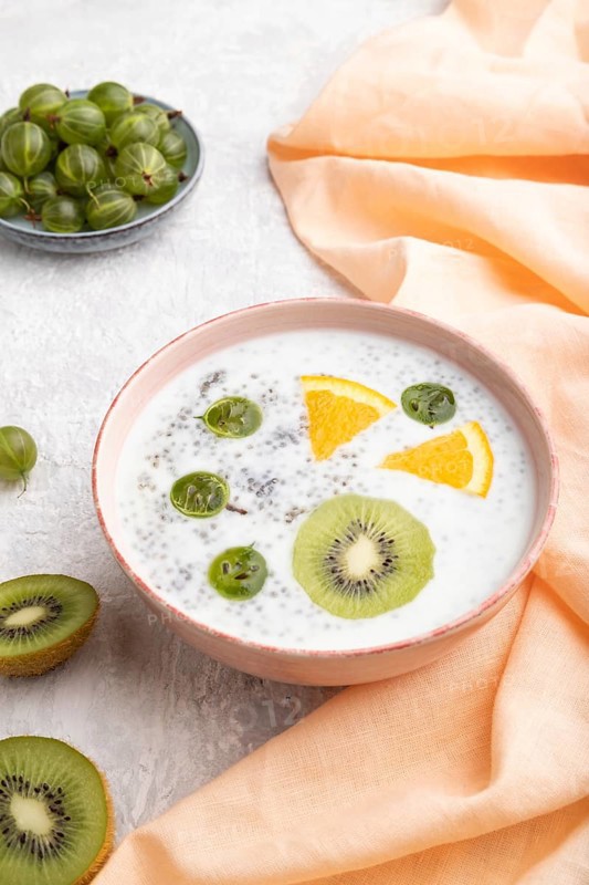 Yogurt with kiwi, gooseberry, chia in ceramic bowl on gray concrete background and orange linen textile. Side view, close up