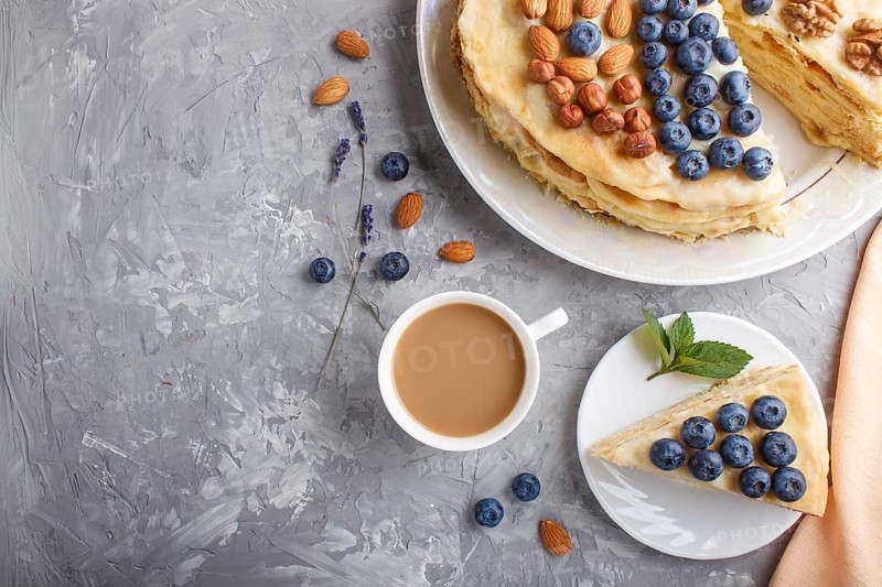 Homemade layered Napoleon cake with milk cream. Decorated with blueberry, almonds, walnuts, hazelnuts, mint on a gray concrete background and cup of coffee. top view. flat lay, copy space