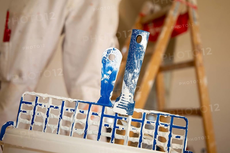 Symbolic image of painting work: Close-up of a paint bucket with scraper grid, in the background a painter's ladder